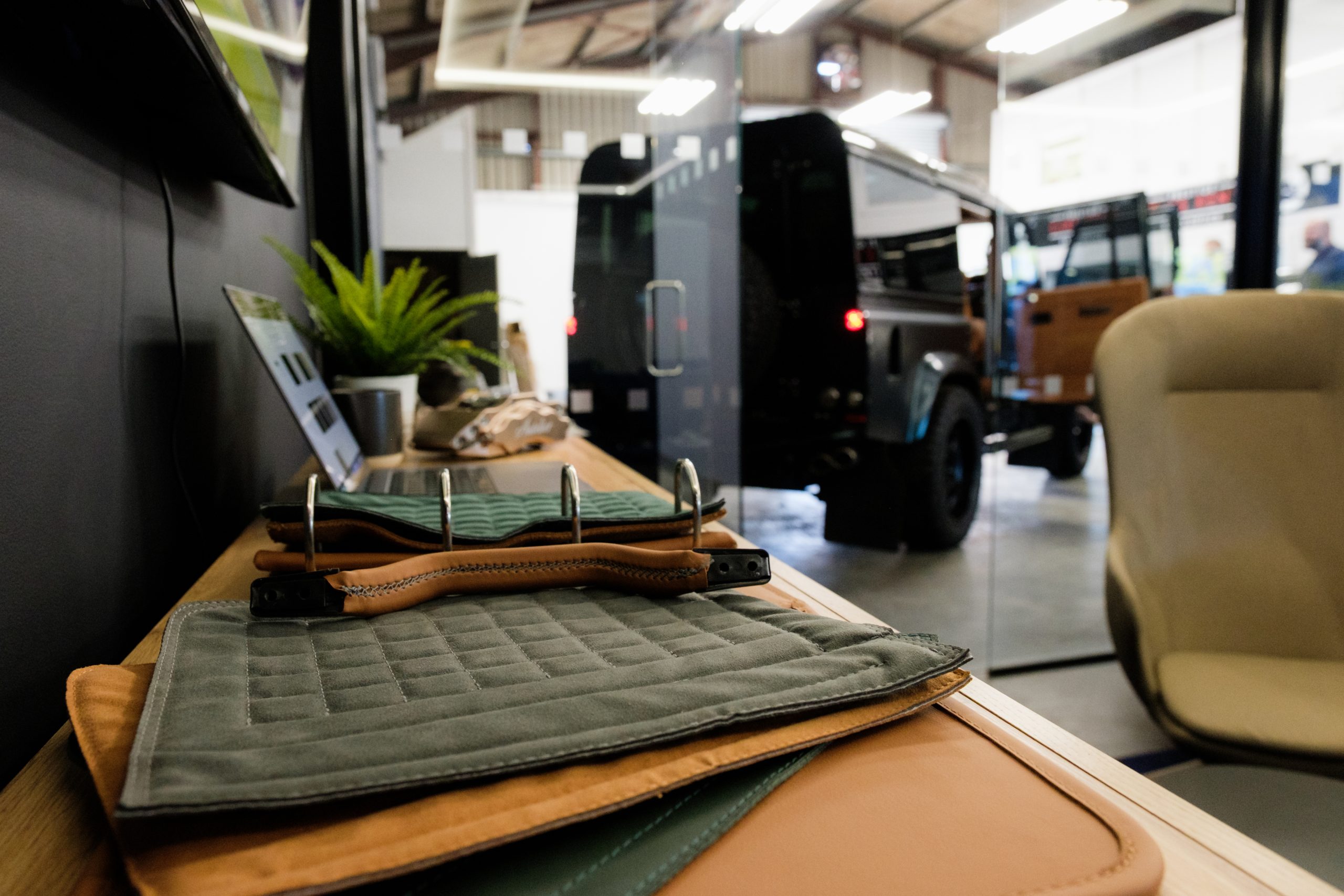 Autelier atelier desk with leather samples, a black Defender parked beyond the glass